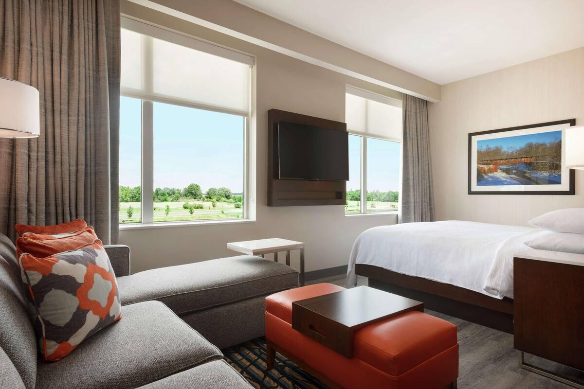 A hotel room with a bed, a sofa, a TV, a small table, and a window with a view of greenery.