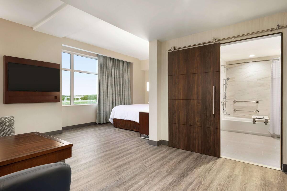 A hotel room with a bed, a desk, a TV, a large wardrobe, and a shower area.