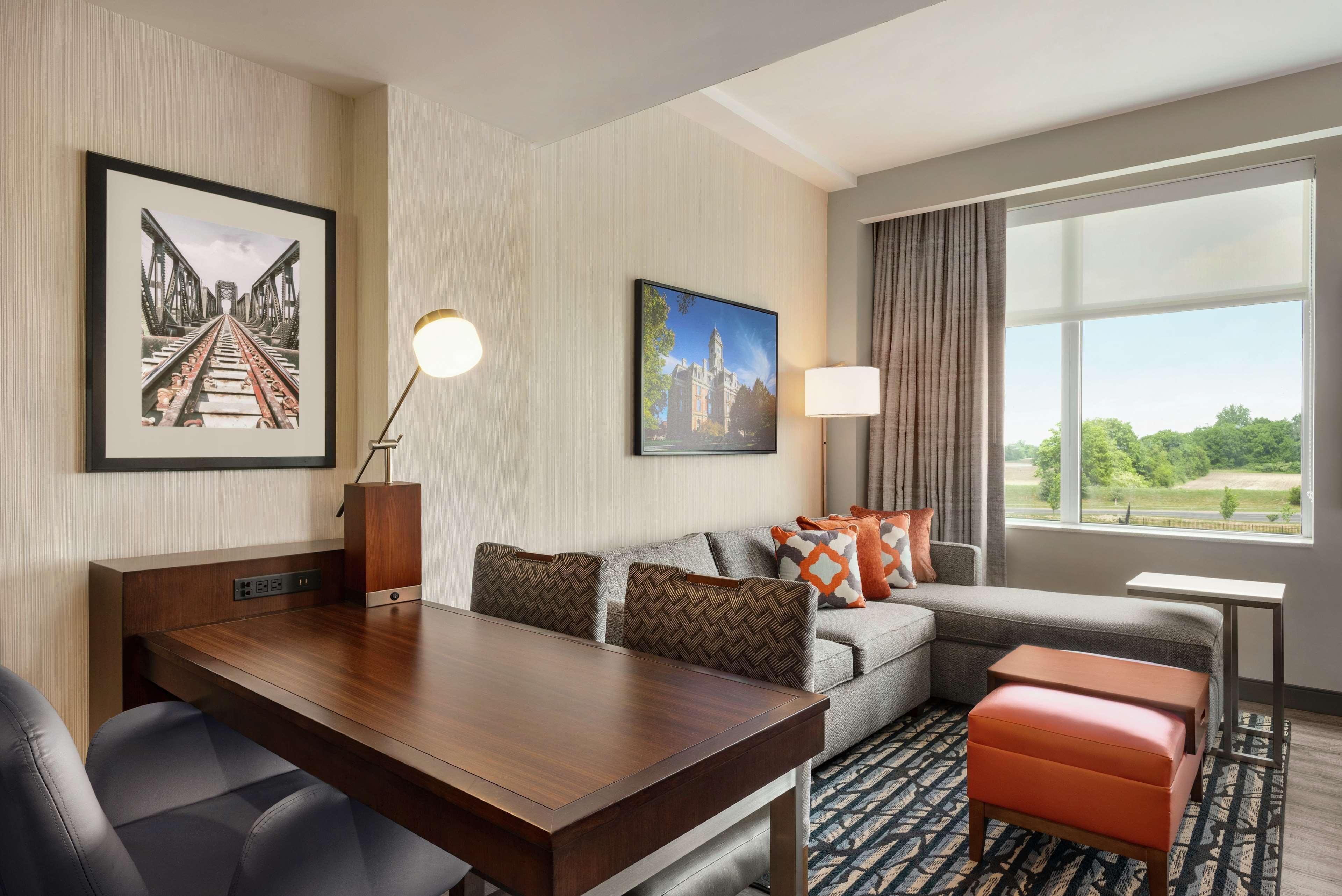 A hotel room with a dining table, a sofa with patterned cushions, a window with a view, and artwork on the walls.