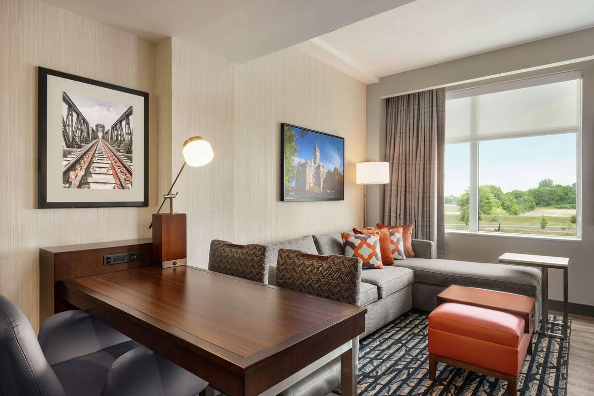 A hotel room with a dining table, a sofa with patterned cushions, a window with a view, and artwork on the walls.