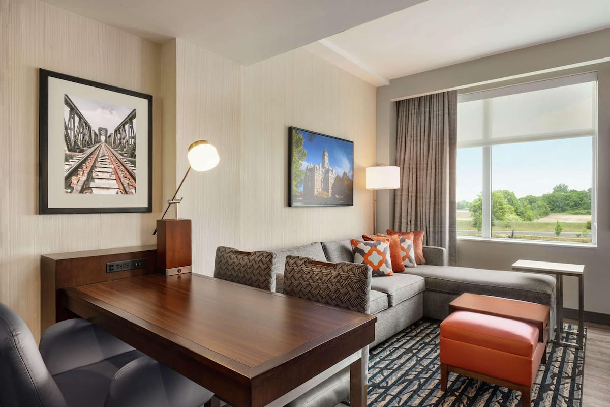 A hotel room with a dining table, a sofa with patterned cushions, a window with a view, and artwork on the walls.