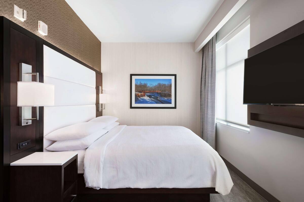 A hotel room with a bed, bedside table, lamp, and a framed picture on the wall.