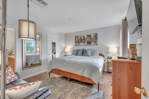Quarto com cama, mesas de cabeceira, escrivaninha, cadeira, janela e um quadro na parede.