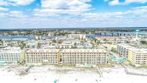 Un paisaje urbano costero con un hotel destacado de varios pisos y balcones, una zona de playa con un paseo marítimo y un río con puentes al fondo.