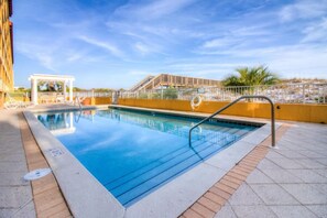 Una piscina con agua cristalina azul, rodeada por un borde de baldosas y una barandilla de metal.