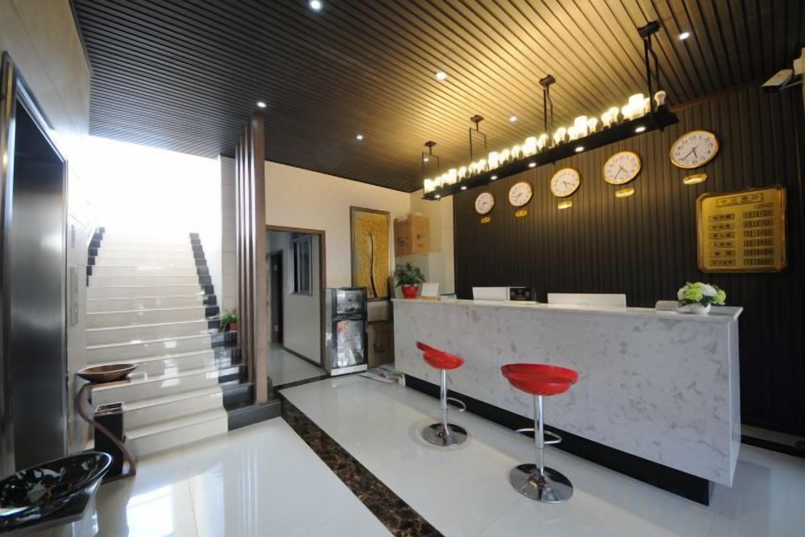 A hotel reception area with a modern design, featuring a reception desk, red bar stools, a staircase, and a wall-mounted clock.