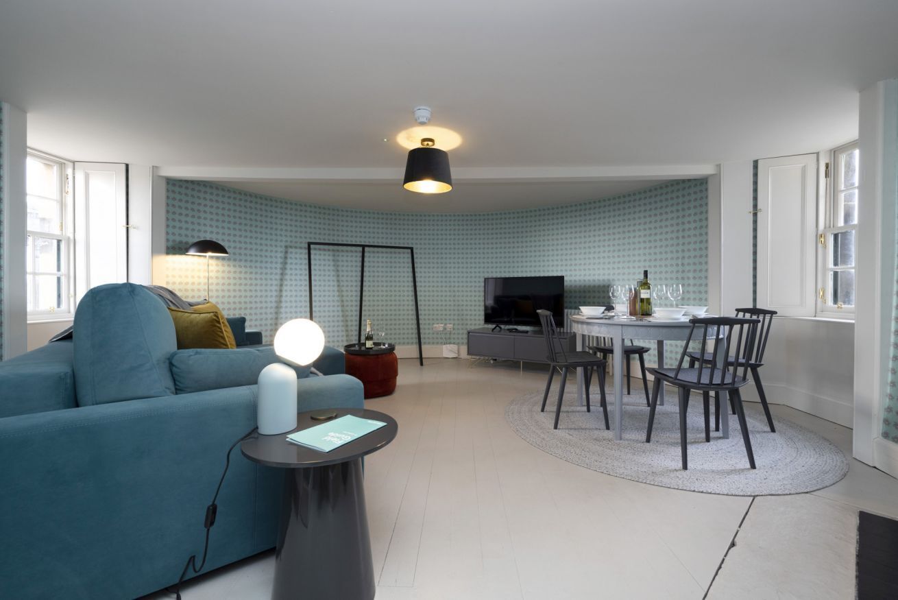 A modern living room with a blue sofa, a round coffee table, a dining table with chairs, a TV, and patterned wallpaper.