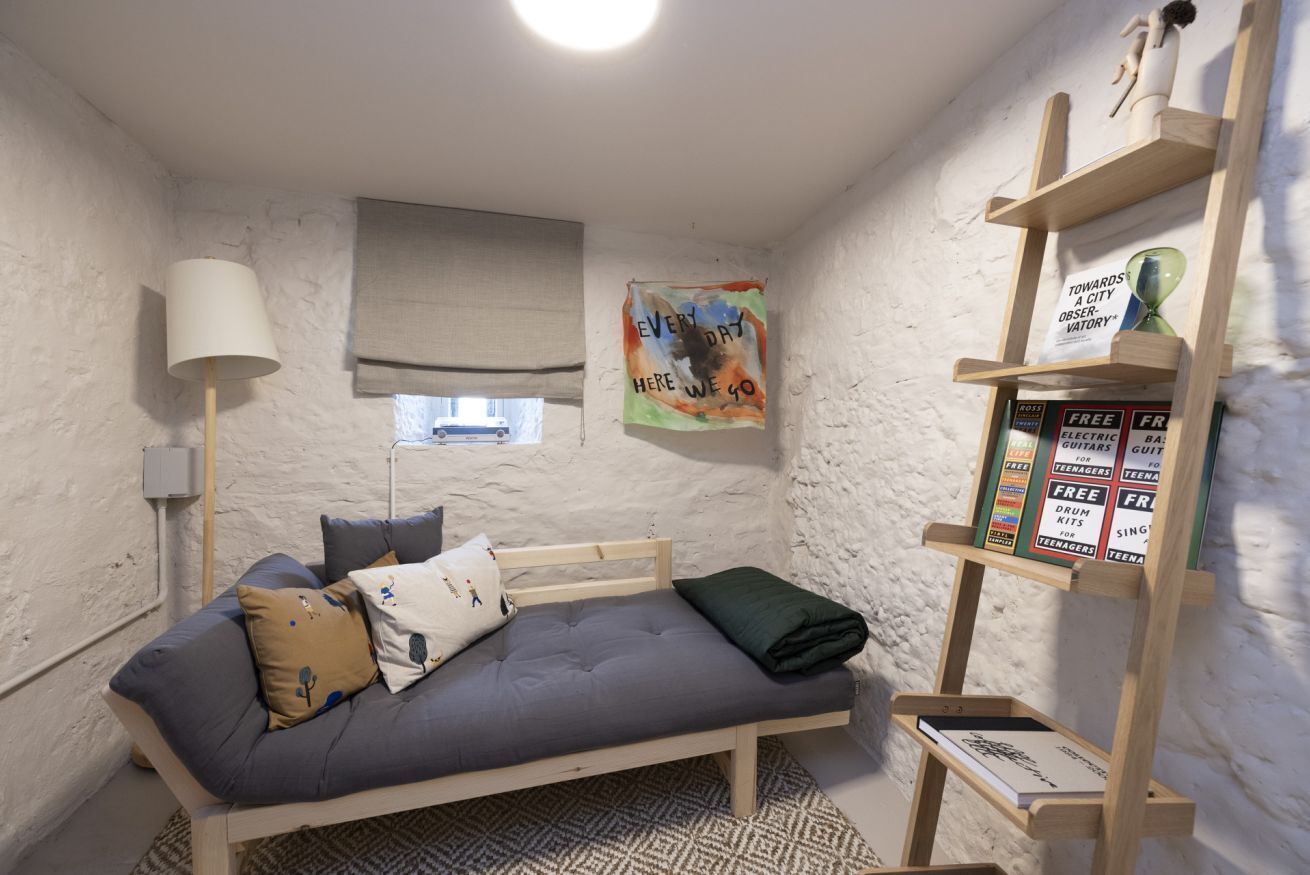 A small, cozy room with a single bed, a lamp, a window with a blind, a poster on the wall, and a shelf with books and a sign.