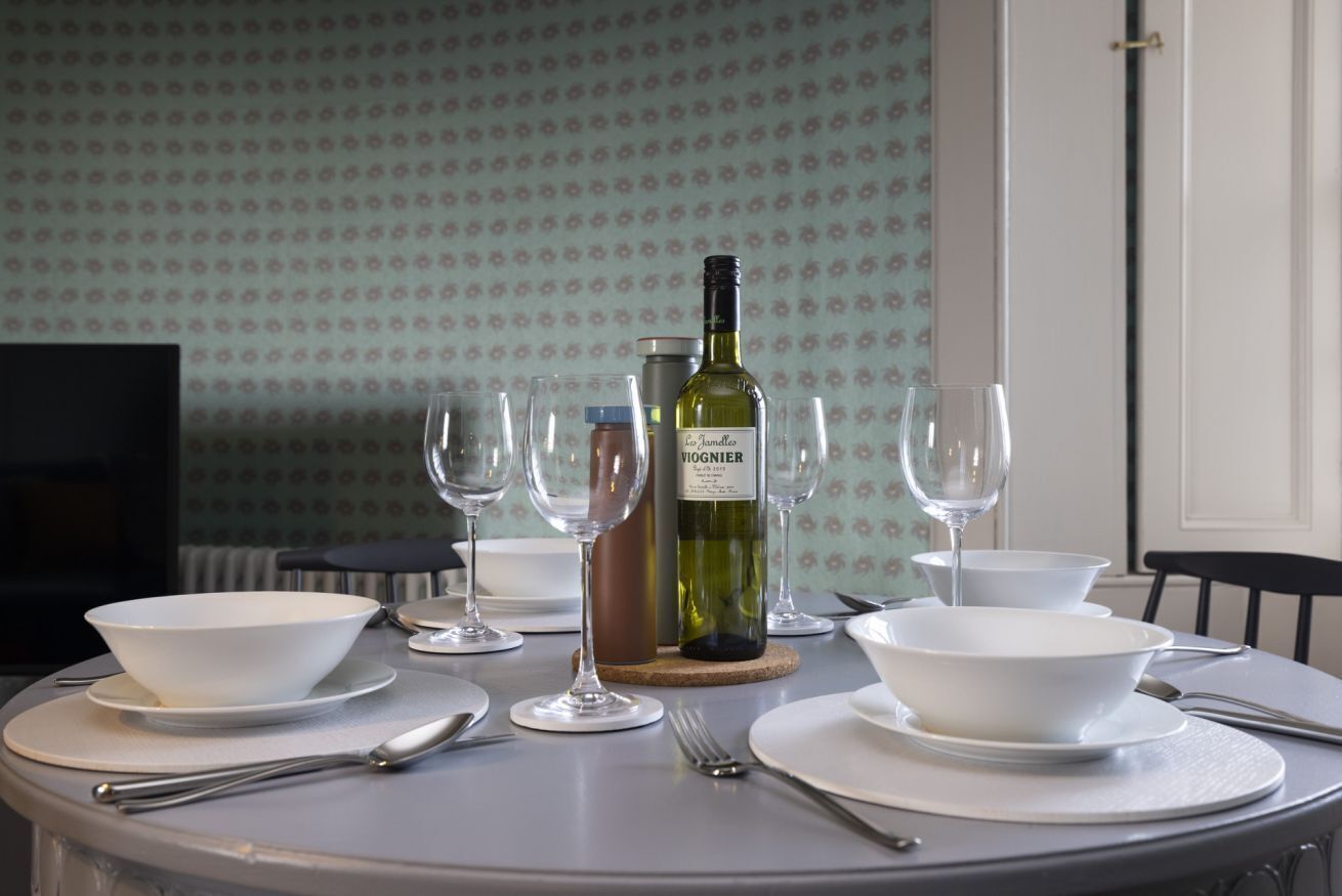 A dining table set with white bowls, plates, cutlery, and wine glasses, with a bottle of wine and a thermos on the table.