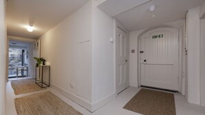 A hallway with white walls, a white door with a green exit sign, and a small table with a plant. - Observatory Apartment at Calton Hill (Edinburgh)