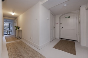 A hallway with white walls, a white door with a green exit sign, and a small table with a plant. - Observatory Apartment at Calton Hill (Edinburgh)
