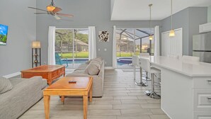 Amplia sala de estar con sofá, mesa de centro y ventilador de techo. Se ve una ventana grande con vista a una piscina y un jardín trasero.