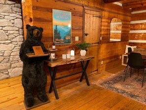 A rustic cabin interior with a stone wall, wooden walls, a wooden table with a bear figurine, a poster of Blue Ridge Mountains, and a chair. - Rustic Elegance, Theatre, Arcade, Sauna, and Mountain Views @ Beaver Cabin (Weaverville)