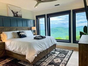 Un dormitorio con cama, mesita de noche, lámpara, ventana grande y ventilador de techo.