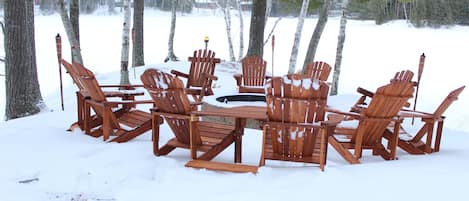 A wooden outdoor seating area with chairs and a central table, surrounded by snow and trees.
