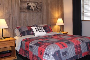 Un lit recouvert d’une couette rouge et grise ornée de motifs animaliers, flanqué de deux tables de chevet avec des lampes, doté d’une tête de lit en bois et agrémenté d’une décoration murale sur le thème de l’ours.