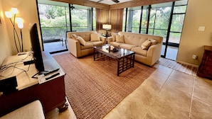 Un salon spacieux avec un canapé beige, une table basse et un téléviseur posé sur un meuble en bois. De grandes fenêtres offrent une vue sur la verdure environnante.