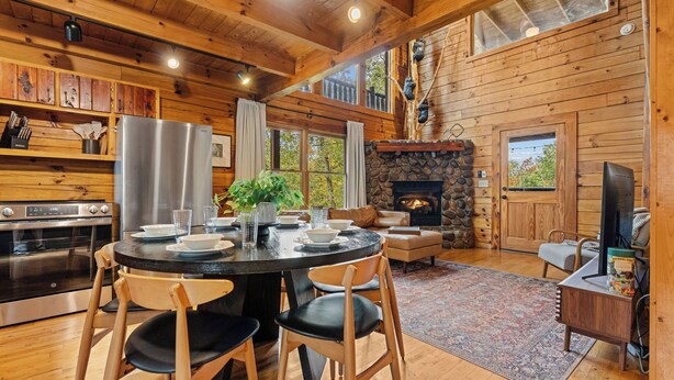 A wooden cabin interior with a dining area, kitchen, fireplace, and a stone fireplace wall. - Scenic Mtn View Cabin with Hot Tub (Seveirville)