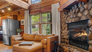 A cozy living room with a stone fireplace, leather sofa, and a large window overlooking greenery. - Scenic Mtn View Cabin with Hot Tub (Seveirville)