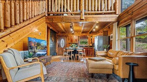 A cozy cabin interior with wooden walls, a dining area, a living room with a sofa and chairs, and large windows offering a view of the outdoors. - Scenic Mtn View Cabin with Hot Tub (Seveirville)