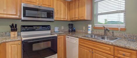 A kitchen with wooden cabinets, a stainless steel microwave, a stove, a dishwasher, granite countertops, and a window with blinds.