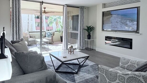 Un salon moderne avec un canapé, une table basse et un fauteuil à motifs. Un téléviseur à écran plat est fixé au mur, et un ventilateur de plafond est visible.