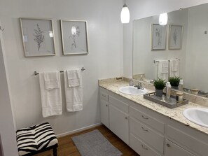 Un bagno moderno con uno specchio grande, due stampe botaniche incorniciate, un lavandino bianco e una panca a righe.