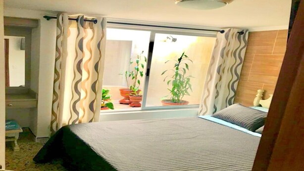Een slaapkamer met een bed, een raam met gordijnen, een kamerplant en uitzicht buiten.