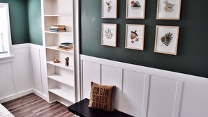 Ein Zimmer mit einem weißen, eingebauten Bücherregal, einer schwarzen Bank mit Kissen sowie gerahmten Pflanzenbildern an der Wand.