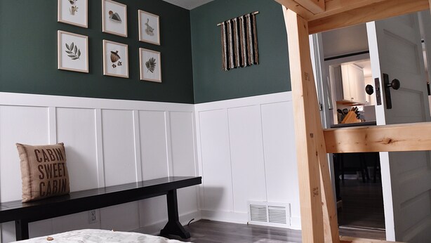 Ein Zimmer mit dunkelgrüner Wand, weißer Wandverkleidung, einem schwarzen Tisch, einem weißen Bett und einer Holzbank.