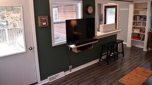 Ein Wohnzimmer mit an der Wand montiertem Fernseher, einem Fenster, einem Bücherregal und einer Tür.