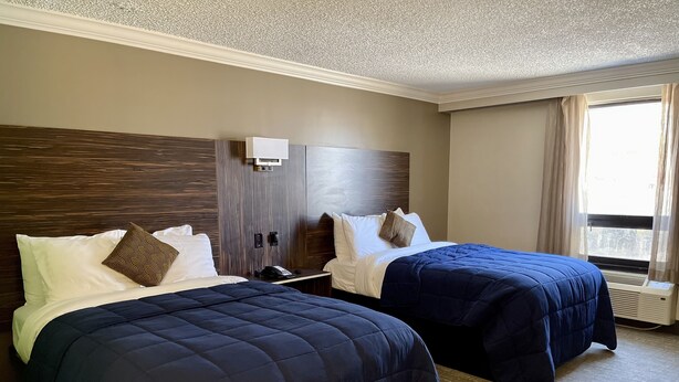 A hotel room with two beds, a wooden headboard, a bedside table with a phone, and a window with curtains.