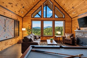 A wooden cabin interior with a pool table, leather sofas, a fireplace, and large windows offering mountain views.