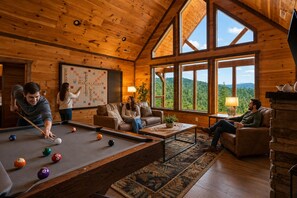 Un intérieur en bois agrémenté d’une table de billard, d’une cheminée et de grandes fenêtres offrant une vue sur les montagnes.