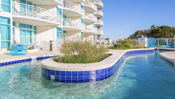 A swimming pool with blue tiles, surrounded by a building with balconies and a garden area. - 1BR Horizons Condo On Beautiful Myrtle Beach Resort w/Amenities (Myrtle Beach)