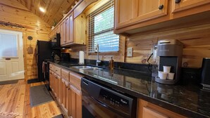 A modern kitchen with wooden cabinets, a black countertop, a dishwasher, a coffee maker, and a window with blinds.