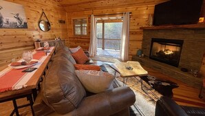 A cozy cabin interior with a wooden interior, a stone fireplace, a sofa, a dining table with glasses, and a television mounted on the wall.