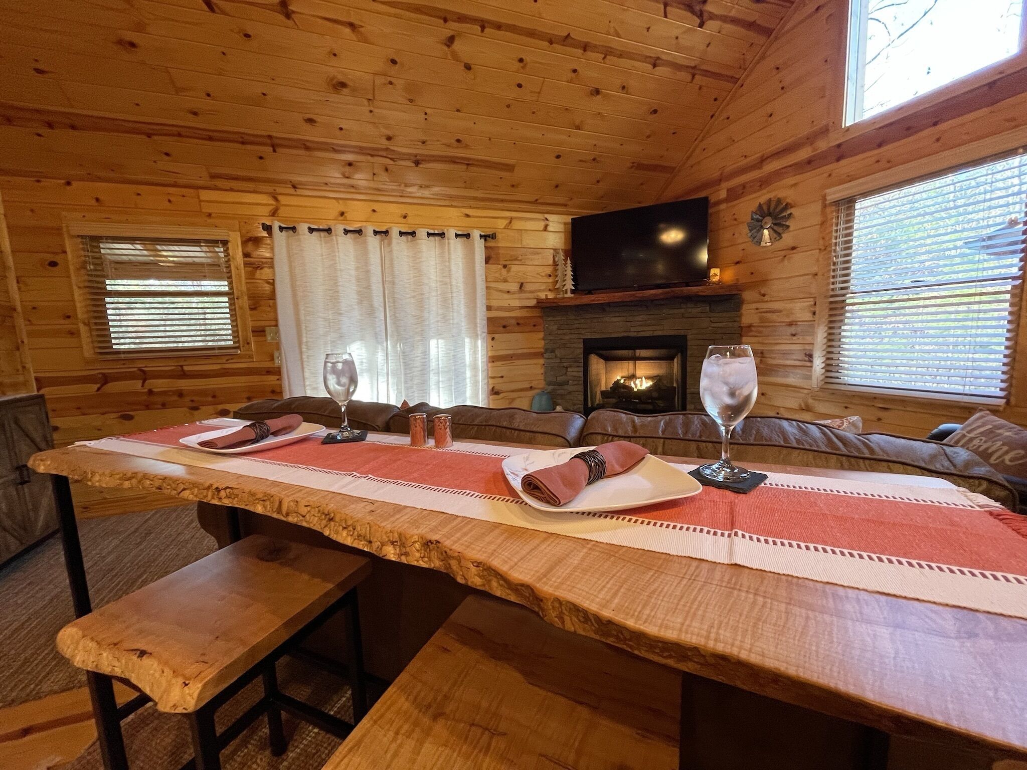 A wooden cabin interior with a long dining table set for two, a fireplace, and a television mounted above it.