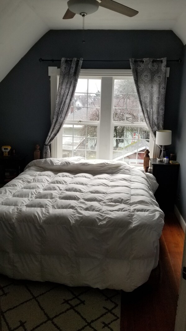 Relax in this serene bedroom with a large bed, a window offering a view of the surrounding area, and a ceiling fan above.