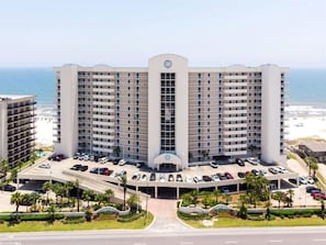 A multi-story hotel with a central entrance, parking area, and a view of the ocean.