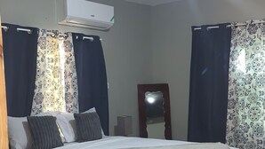 A bedroom with a bed, curtains, an air conditioning unit, a mirror, and a wooden floor.