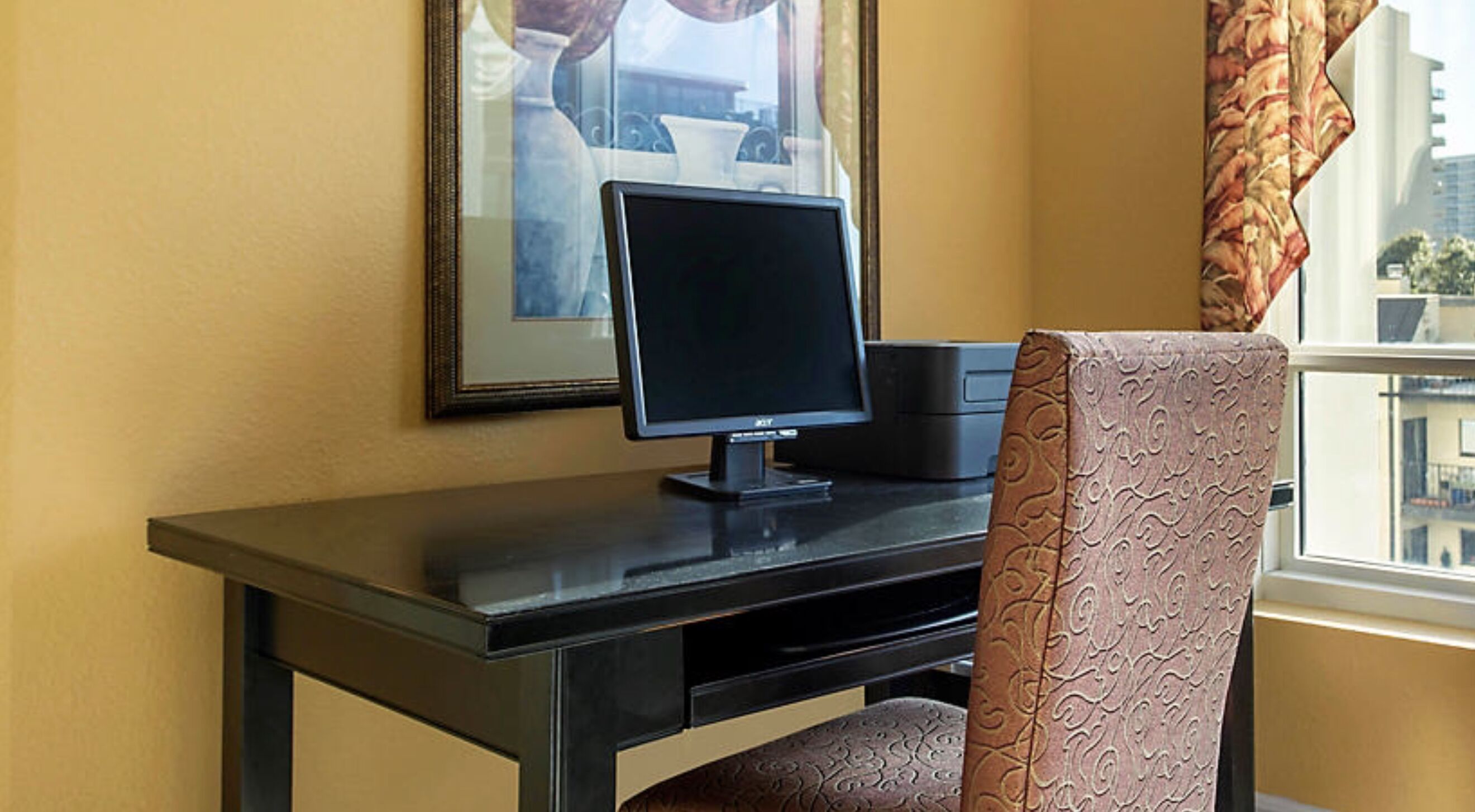 A hotel room with a desk, a computer, a chair, and a window with a view of buildings.