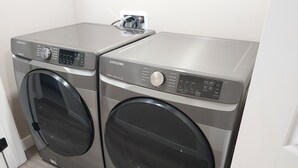 Två Samsung-tvättmaskiner med frontmatning i ett tvättrum med en vit vägg och trägolv.