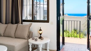 Ein Zimmer mit einer Couch, einem kleinen Tisch mit Dekoration, einem Fenster mit Meerblick und einem Balkon mit Geländer.