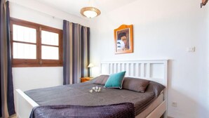 Ein Schlafzimmer mit Bett, Fenster mit Vorhängen, Nachttisch mit Lampe und einem gerahmten Bild an der Wand.