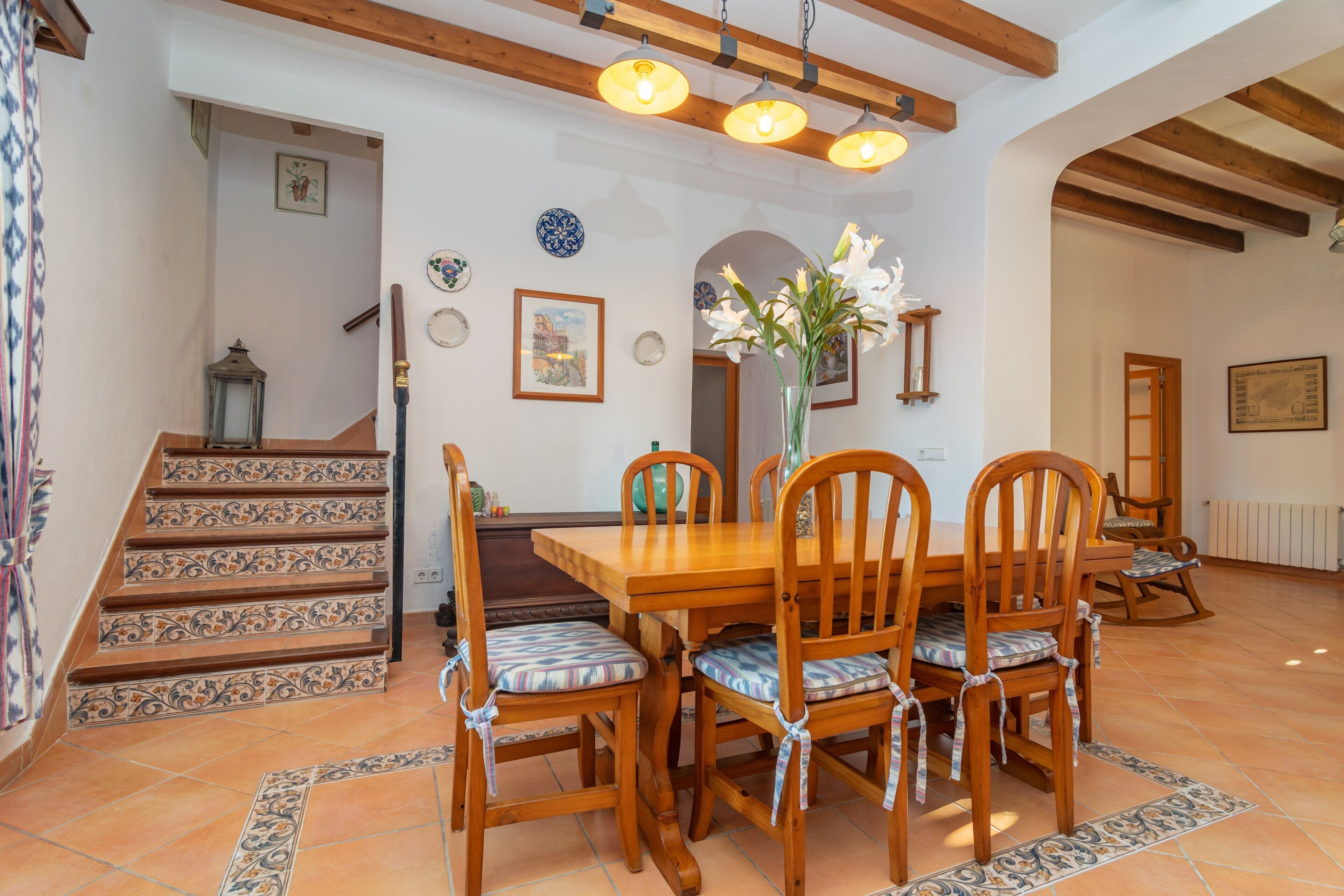 Un coin repas avec une table et des chaises en bois, un escalier orné de carreaux décoratifs et un plafond avec des poutres apparentes.