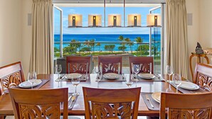 A dining area with a table set for six, overlooking a beach with palm trees and the ocean.