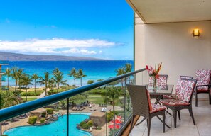 Ein Balkon mit Blick auf Pool und Strand, Korbstühle und ein Tisch mit Blumen in einer Vase.