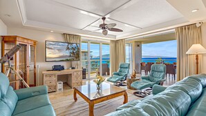 A spacious living room with a large sofa, a coffee table, and a desk with a telescope. There is a large window with a view of the ocean.