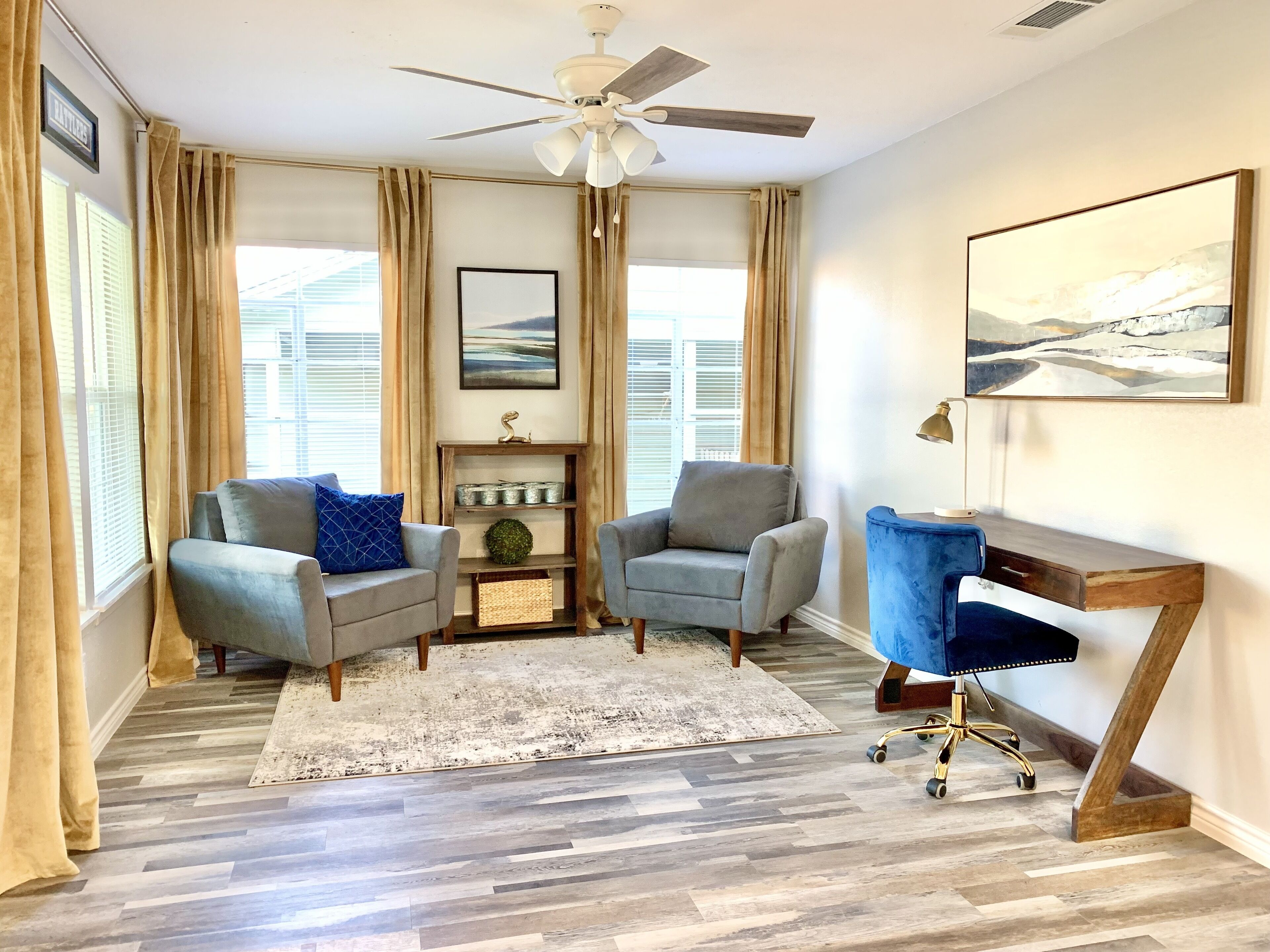 A living room with two grey armchairs, a blue desk, a ceiling fan, and a large window with blinds.